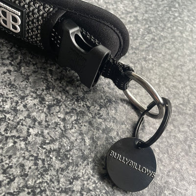 Close‑up of BullyBillows Combat Collar in black showing metal clip, woven nylon strap, and engraved BullyBillows tag resting on grey textured surface.